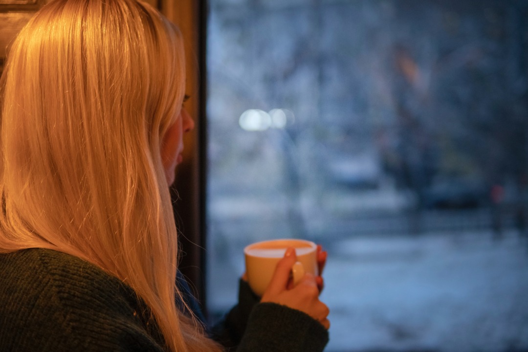 woman drinking from mug and looking out at snowy evening