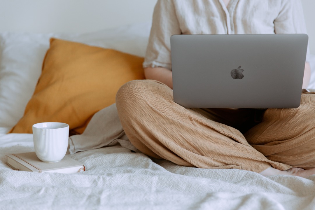 Person sat in bed with computer, mug, and journal